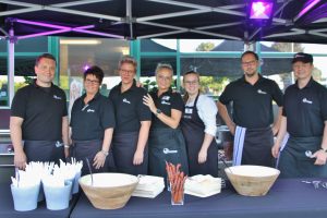 Gruppe von sieben Personen in schwarzen T-Shirts und Schürzen bei einer Grillveranstaltung.