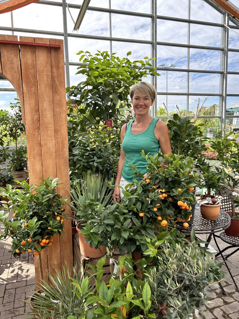 Frau in grünem ärmellosem Oberteil steht in einem Gewächshaus umgeben von mehreren Topfpflanzen mit kleinen orangen Früchten