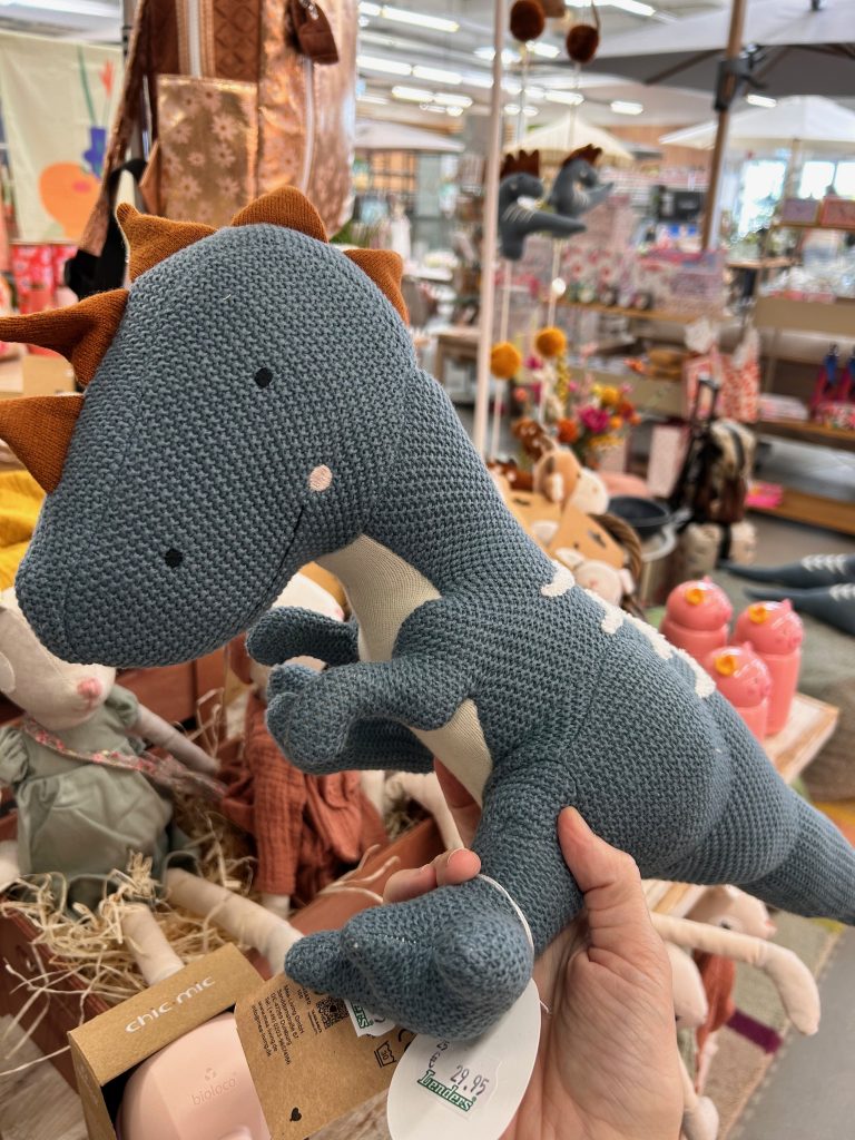 Hand hält gestricktes Dinosaurier-Spielzeug in einem Laden mit weiteren Spielwaren im Hintergrund