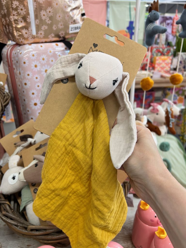 Hand hält gelbes Schmusetuch mit Hasenkopf aus Stoff in einem Laden mit Babyartikeln