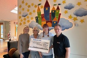 Vier Personen stehen vor einer Wand mit einer bunten Burg- und Sternenwandmalerei und halten einen großen Scheck der Gladbacher Bank über 1400 Euro.