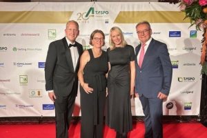 Zwei Frauen in schwarzen Abendkleidern und zwei Männer in Anzügen stehen auf rotem Teppich vor einer Sponsorenwand mit Logos und Texten.