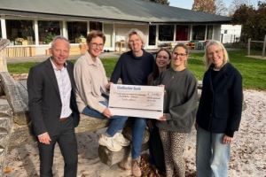 Fünf Personen stehen und sitzen auf einem Holzspielgerät im Sand, eine Person hält einen großen Scheck mit dem Logo der Volksbank.